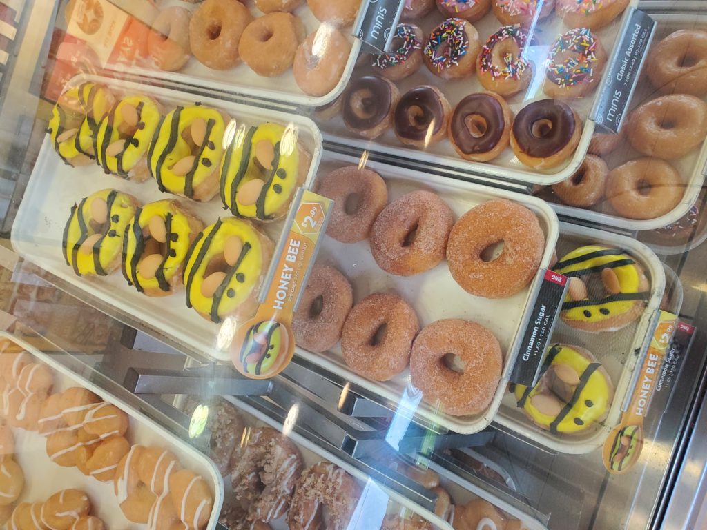 Pans of honey themed donuts in glass display case. Mixture of cake and fluffy type doughnuts